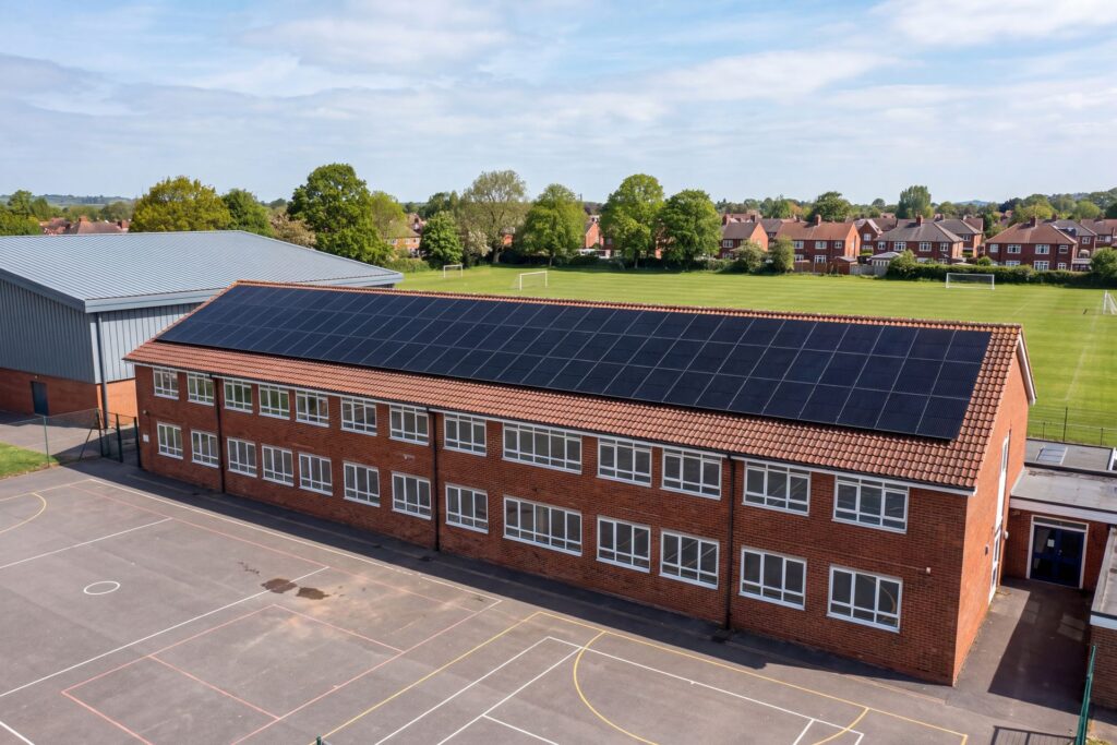 Aerial view of a secondary school classroom block with all-black solar panels covering its roof, beside the sports hall and playing fields